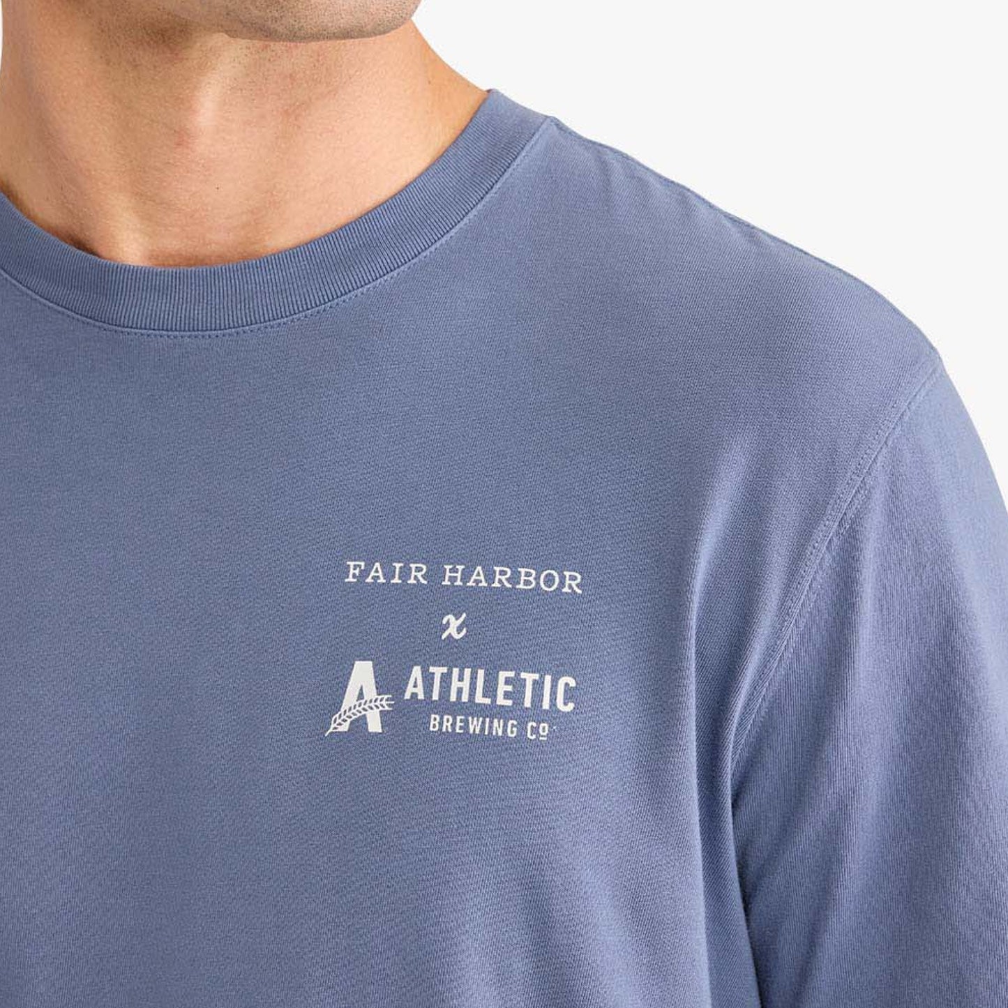 Close-up detail of a faded blue T-shirt showing the white ‘Fair Harbor x Athletic Brewing Co.’ logo printed on the chest.