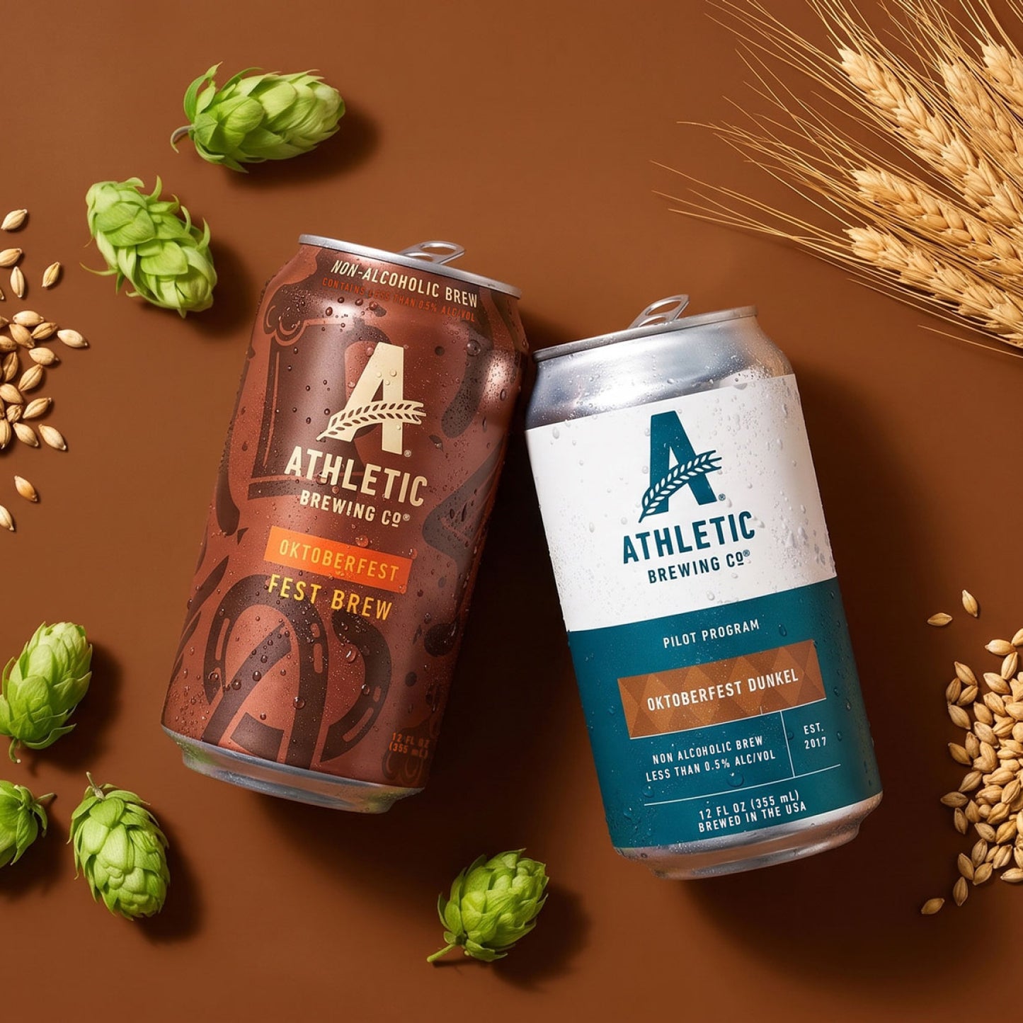 Athletic Brewing Oktoberfest Fest Brew and Oktoberfest Dunkel cans surrounded by green hops, barley grains, and wheat on a warm brown background.