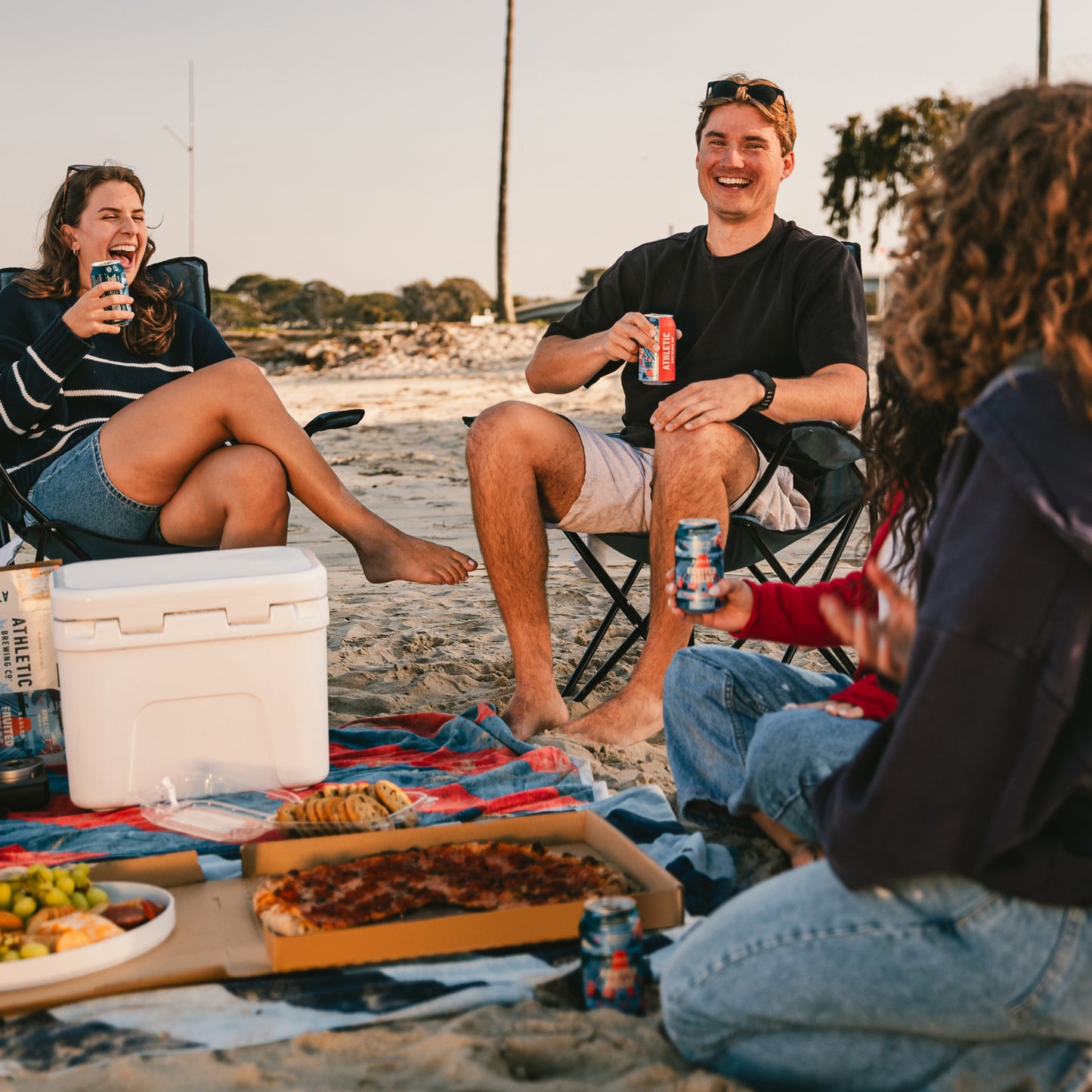 four people sitting on the beach holding cans of Fruited Fields Non alcoholic brew with a cooler and food on a blanket