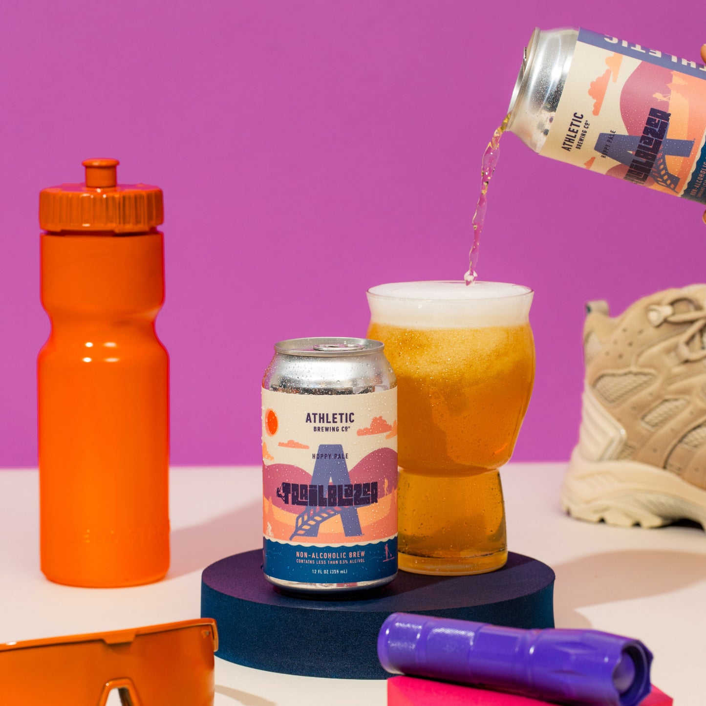 Lifestyle flat lay featuring a Trailblazer can and beer being poured into a glass, surrounded by outdoor gear including an orange water bottle, hiking boot, sunglasses, and a purple flashlight.