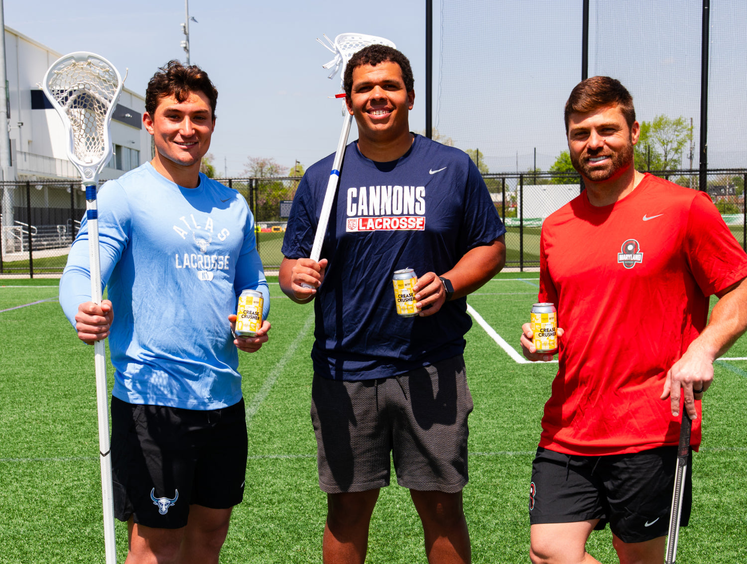 Three professional lacrosse players standing on an artifical turf field holding lacrosse sticks and cans of crease crusher non alcoholic brews