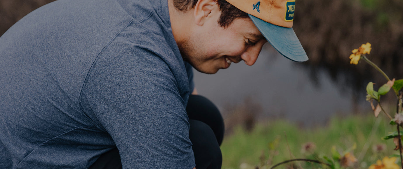 A man in a blue long-sleeve shirt and a tan cap with a blue brim crouches outdoors near yellow flowers.