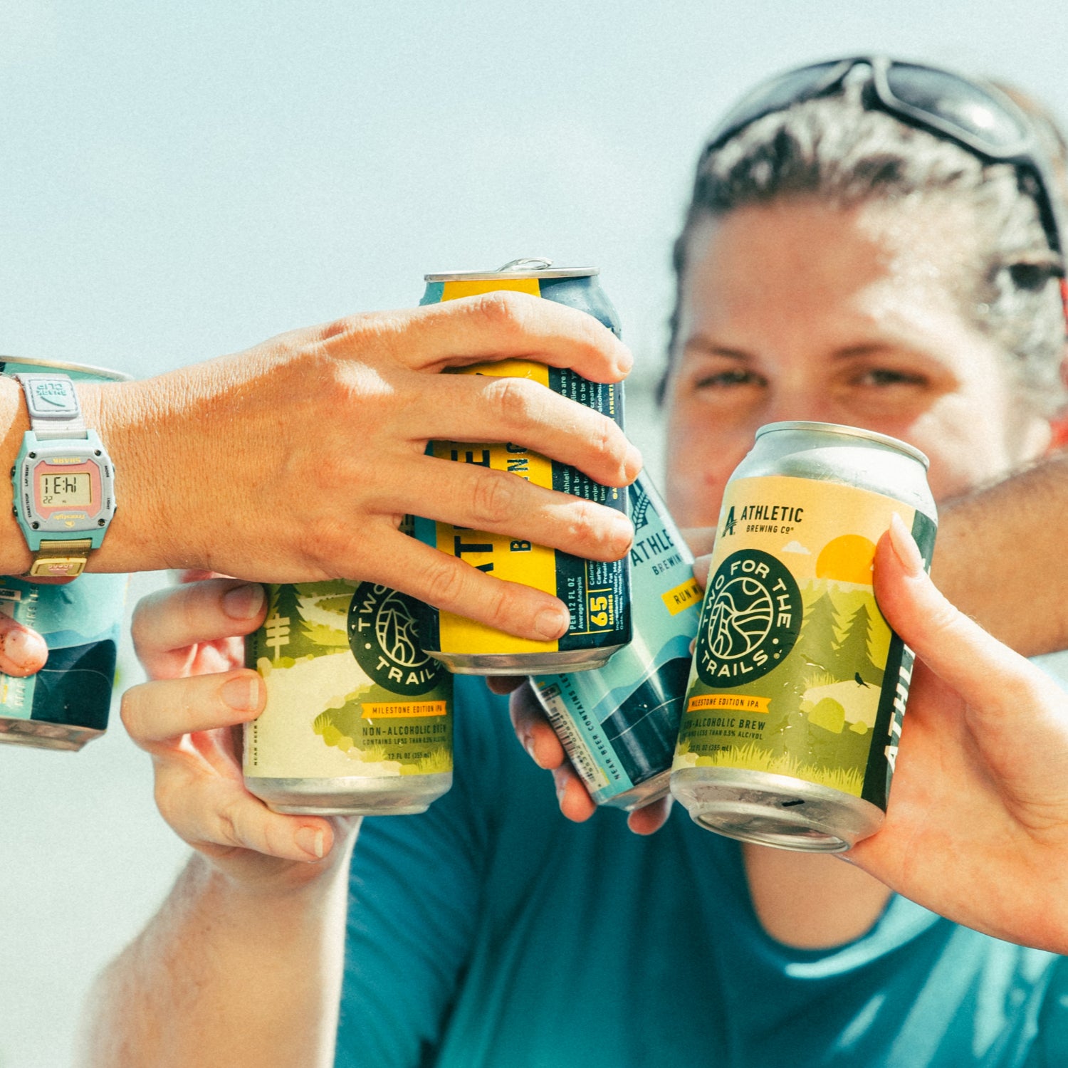 A first-person perspective of four people clinking cans of Two For The Trails and other Athletic Brewing beers in a celebratory outdoor toast.