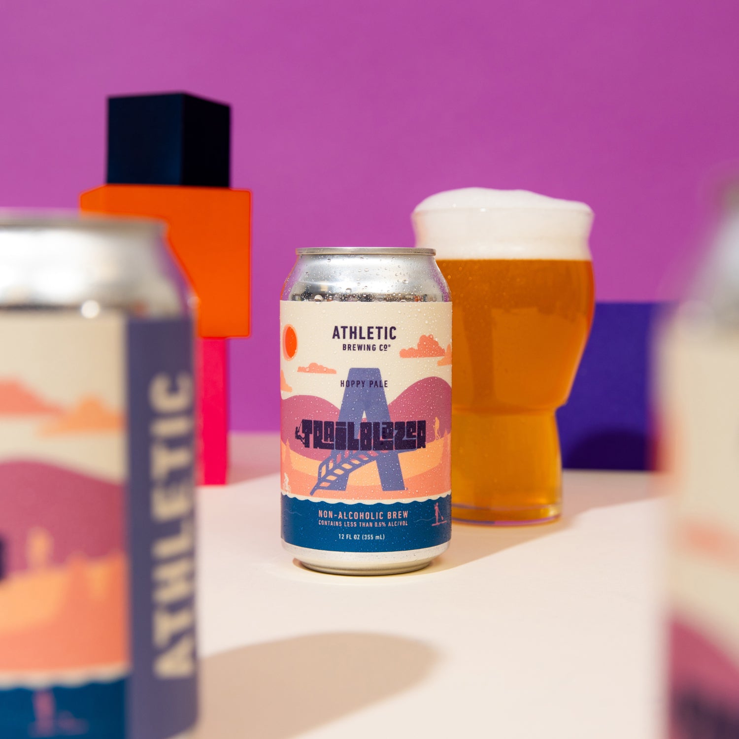 A centered Trailblazer non-alcoholic beer can with a full glass of beer in the soft-focus background, set against a vibrant purple backdrop with geometric orange and blue shapes.