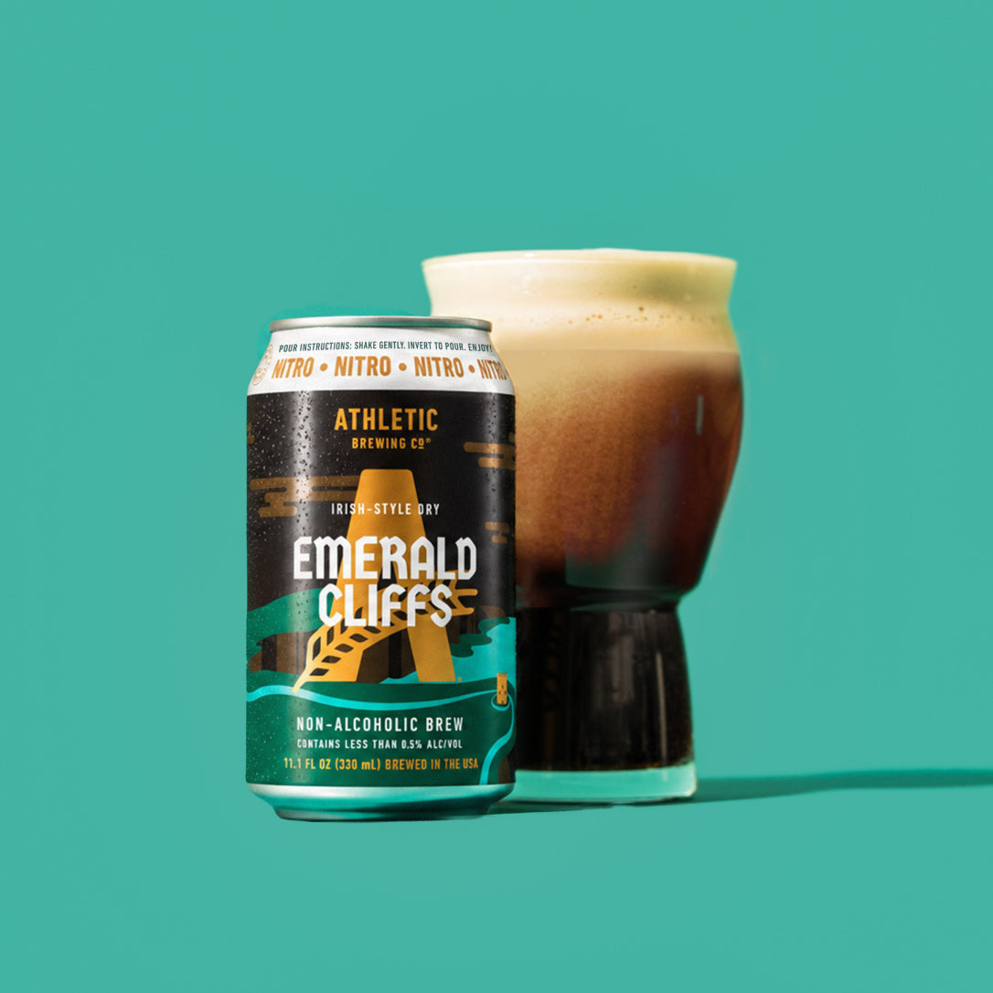 Athletic Brewing Emerald Cliffs non-alcoholic nitro brew can beside a glass of dark beer with a thick, creamy foam head on a teal background.