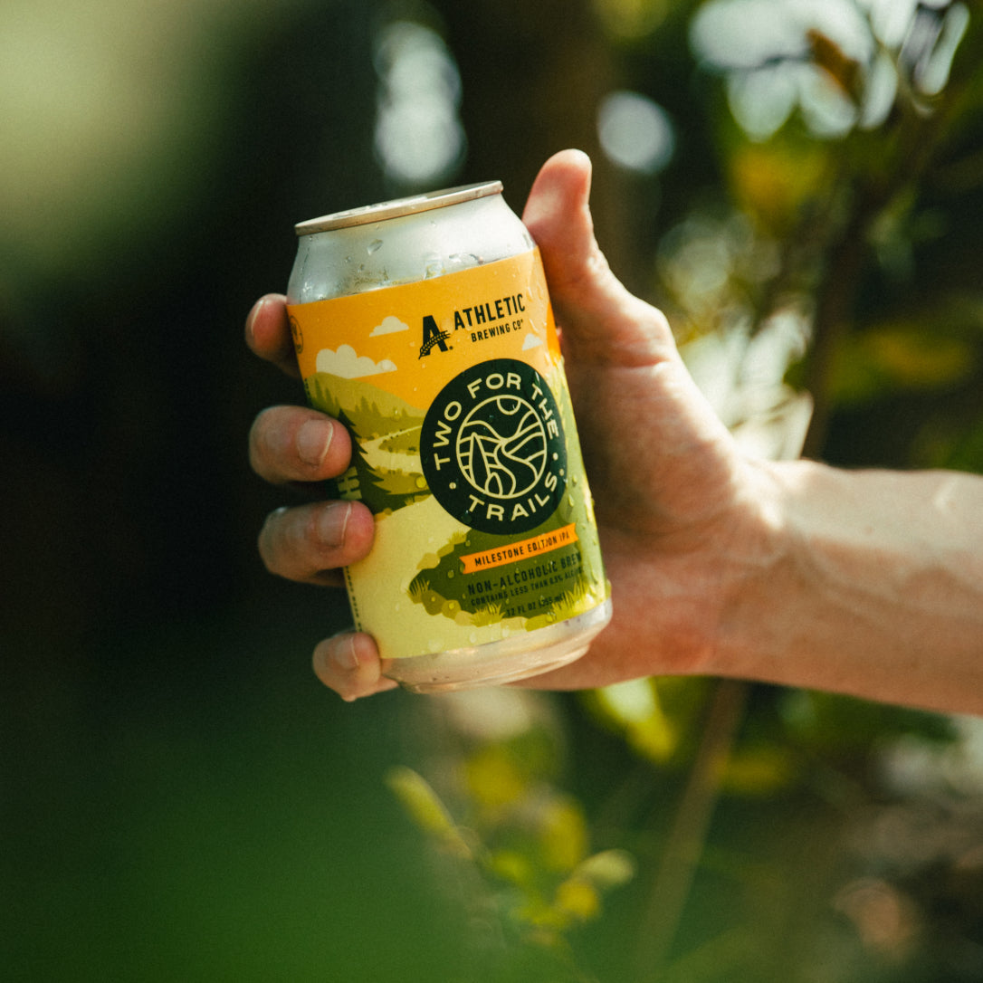 A close-up shot of a hand gripping a cold Two For The Trails IPA can with a blurred green nature background.