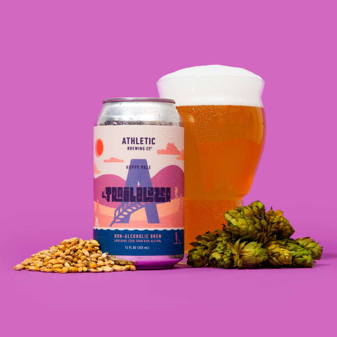 A single can of Trailblazer non-alcoholic beer positioned next to a tall glass of golden ale with a thick white head, flanked by a small pile of malted grain and several fresh green hop cones.