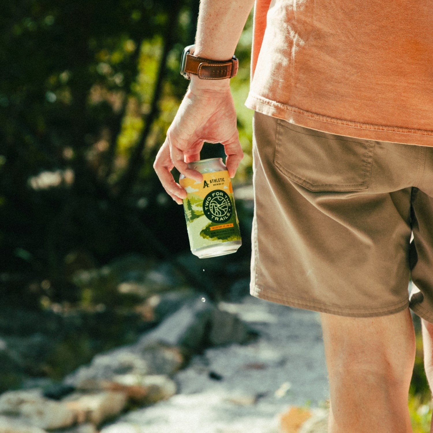 A close-up of a person's hand holding a chilled can of Two For The Trails Milestone Edition IPA while walking on a sunlit trail.