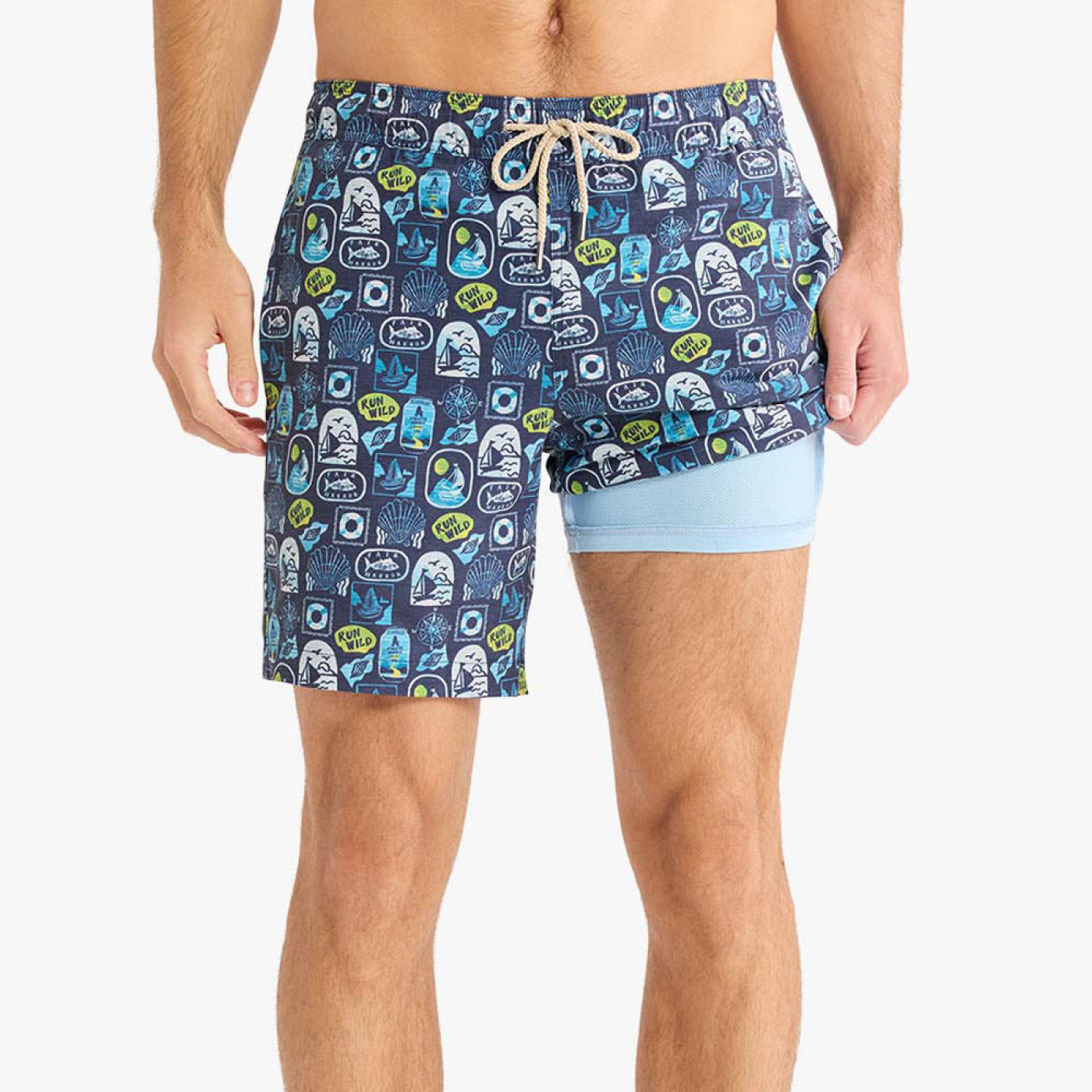 Front view of a person wearing navy blue printed swim shorts with a beige drawstring waistband. One side of the shorts is lifted to reveal a light blue built-in compression liner underneath. The shorts feature an allover coastal-themed pattern with sailboats, shells, and graphic patches.