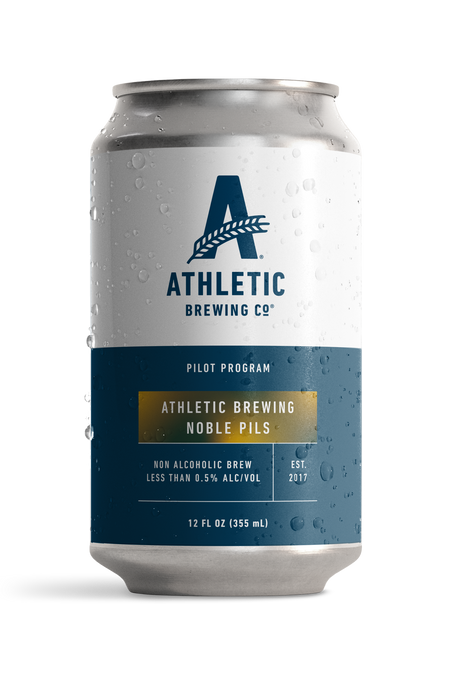 12 fl oz can of Athletic Brewing Pilot Program Noble Pils non-alcoholic beer. The silver can features a white upper label with the Athletic Brewing Co. logo and a navy blue lower label with gold accents displaying the beer name, “Noble Pils.” Additional text notes it is a non-alcoholic brew with less than 0.5% ABV. Condensation droplets cover the can, shown against a black background.