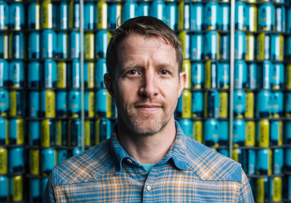 Athletic Brewing Company brewer wearing a blue plaid shirt, standing in front of stacked blue and yellow Athletic Brewing non-alcoholic beer cans at the brewery.