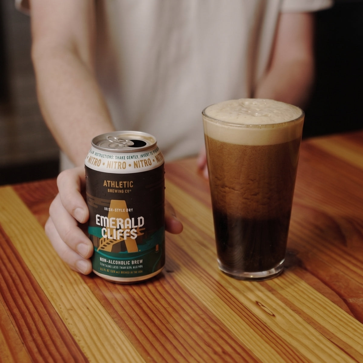 Alt text: Athletic Brewing Emerald Cliffs Irish-Style Dry non-alcoholic nitro brew can beside a glass of dark beer with a thick, creamy foam head on a wooden table.