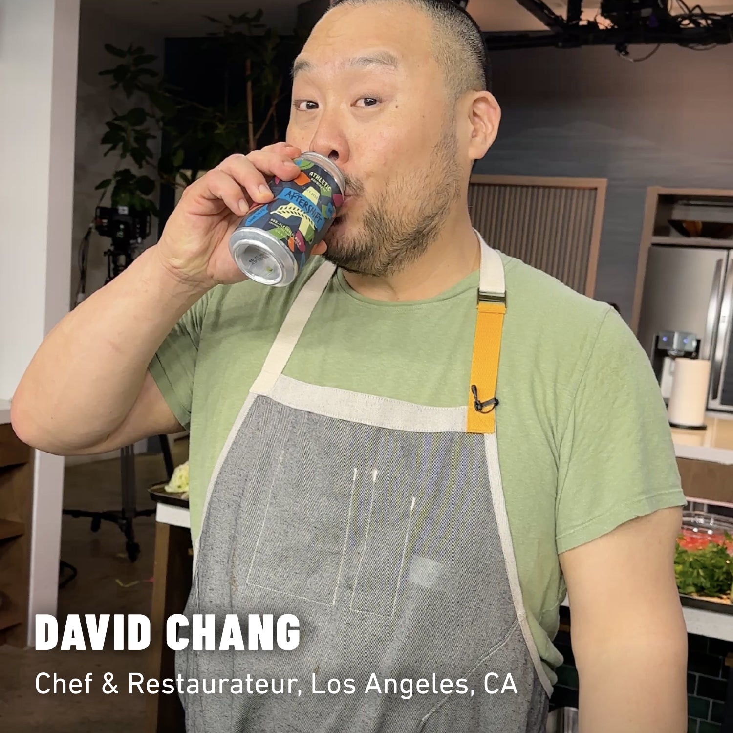 David Chang in a green shirt and grey chef apron holding a can of non-alcoholic beer