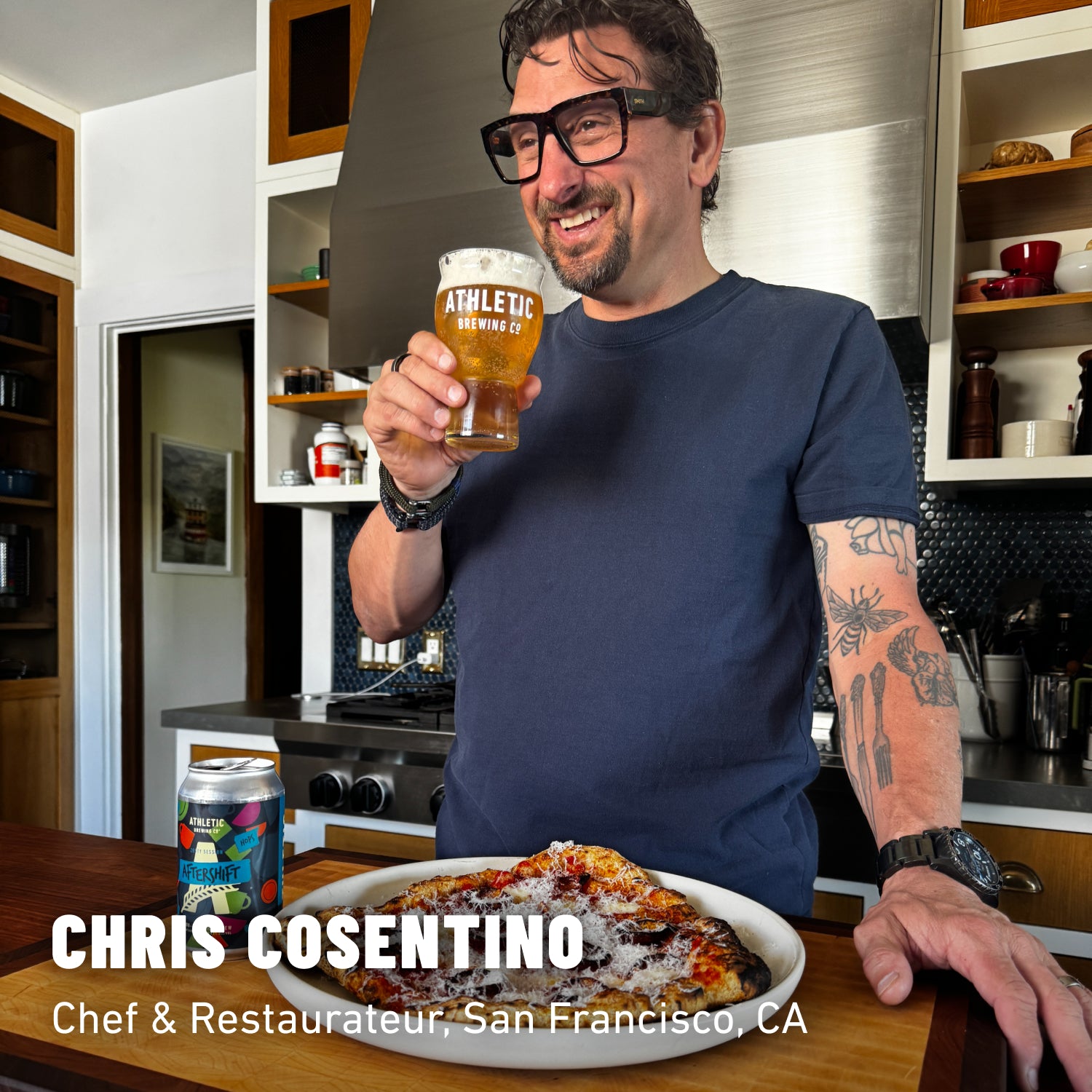 Chris Cosentino in a blue shirt holding a glass of non-alcoholic beer standing over a pizza