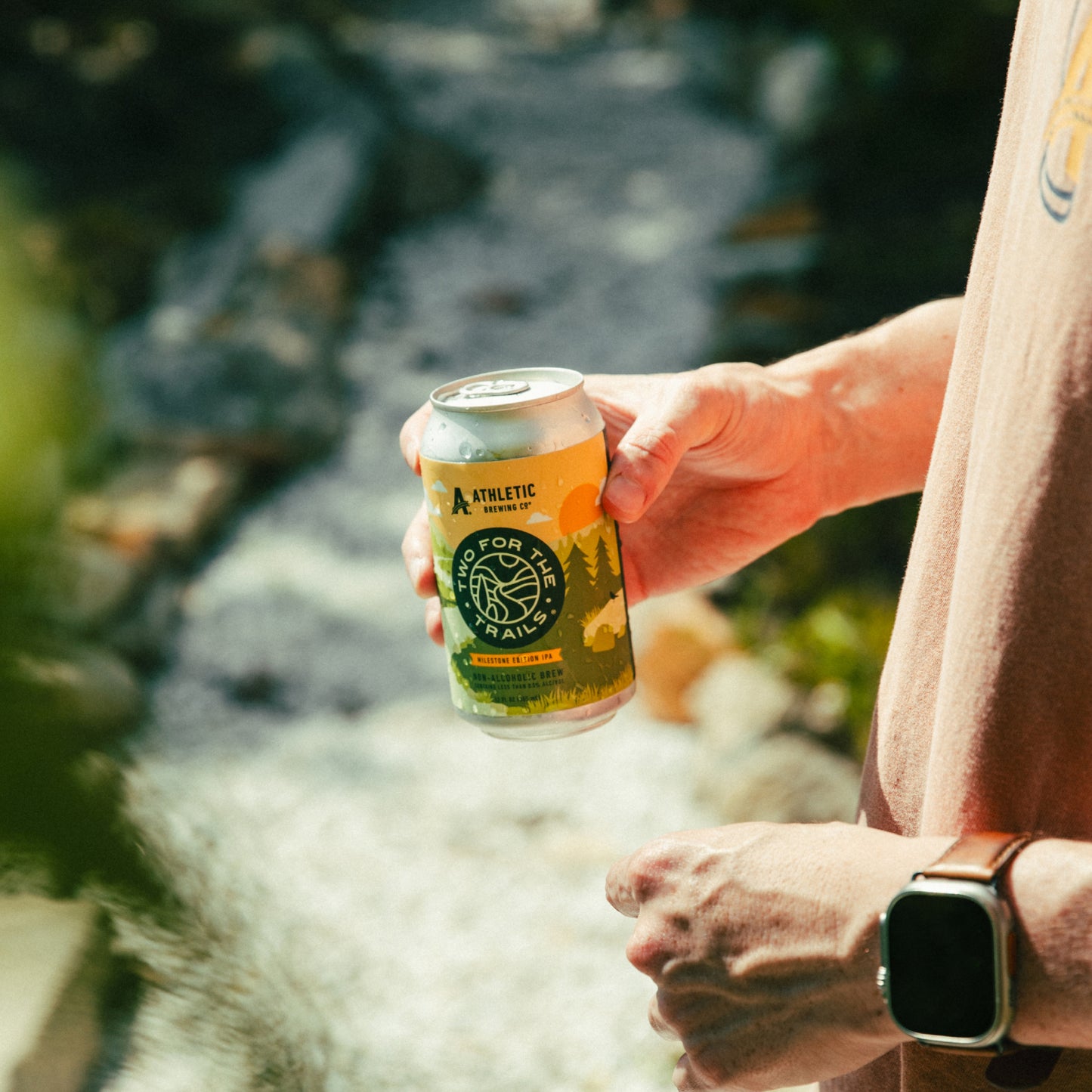A person on a hike holding a can of Two For The Trails Milestone Edition IPA near a rocky stream.