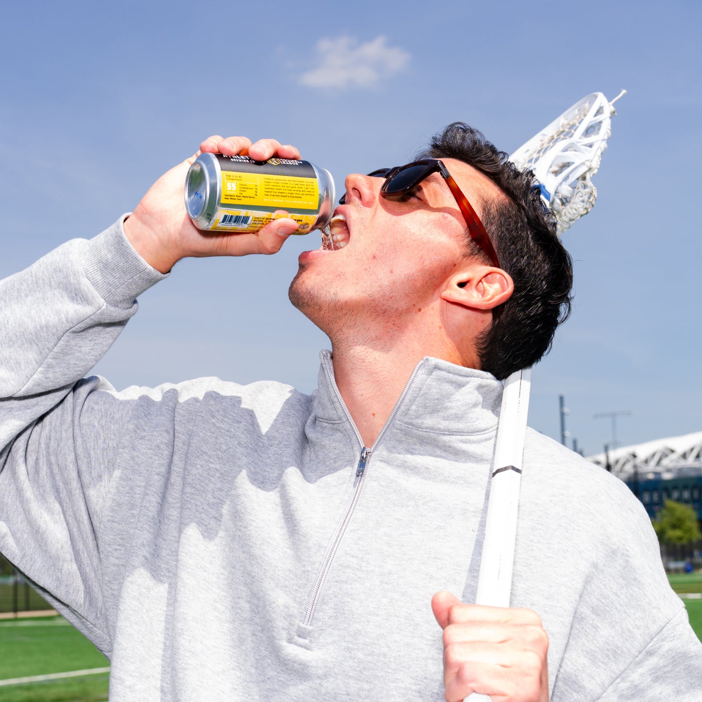 Person with grey quarter zip wearing sunglasses holding a lacrosse stick pouring a can of non alcoholic beer in their mouth