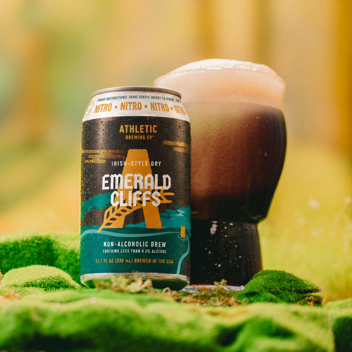 Athletic Brewing Emerald Cliffs non-alcoholic nitro brew can beside a glass of dark beer with a creamy foam head in a mossy outdoor setting.