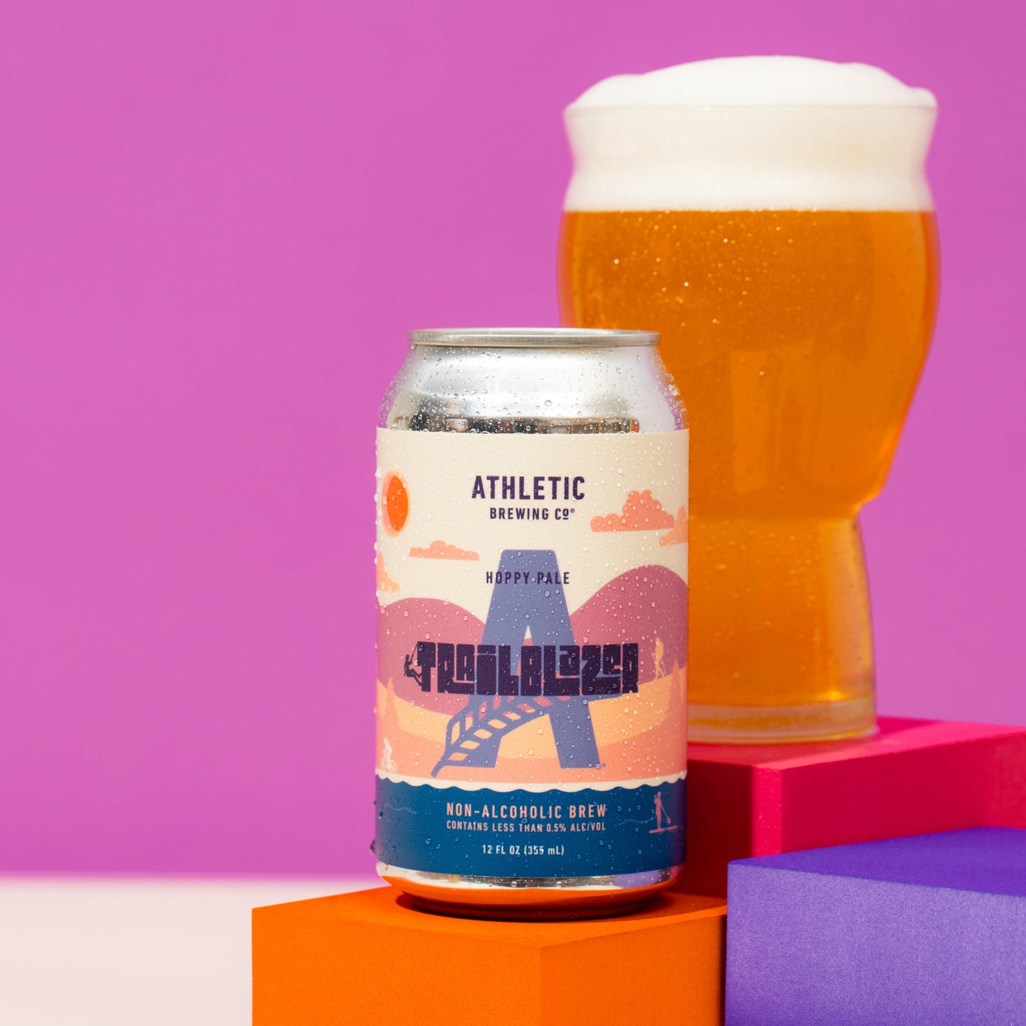 A can of Trailblazer non-alcoholic beer resting on colorful orange and purple geometric blocks with a full, frothy glass of golden beer behind it against a purple background.