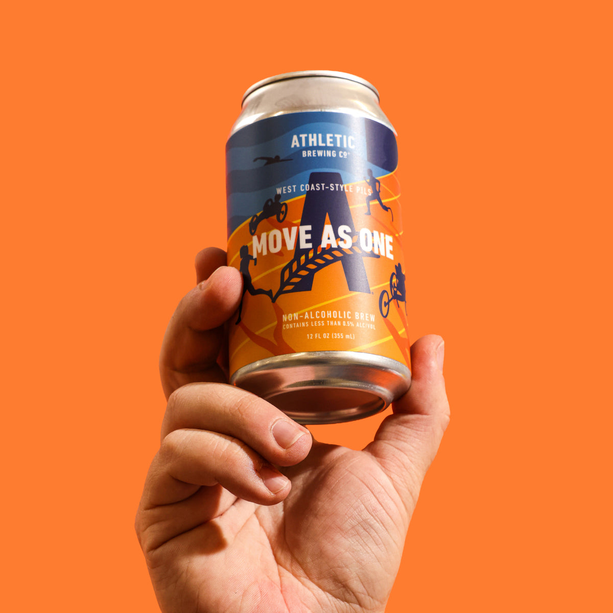 Close-up of a hand holding an Athletic Brewing Move As One West Coast-Style Pils non-alcoholic beer can against a vibrant orange background.