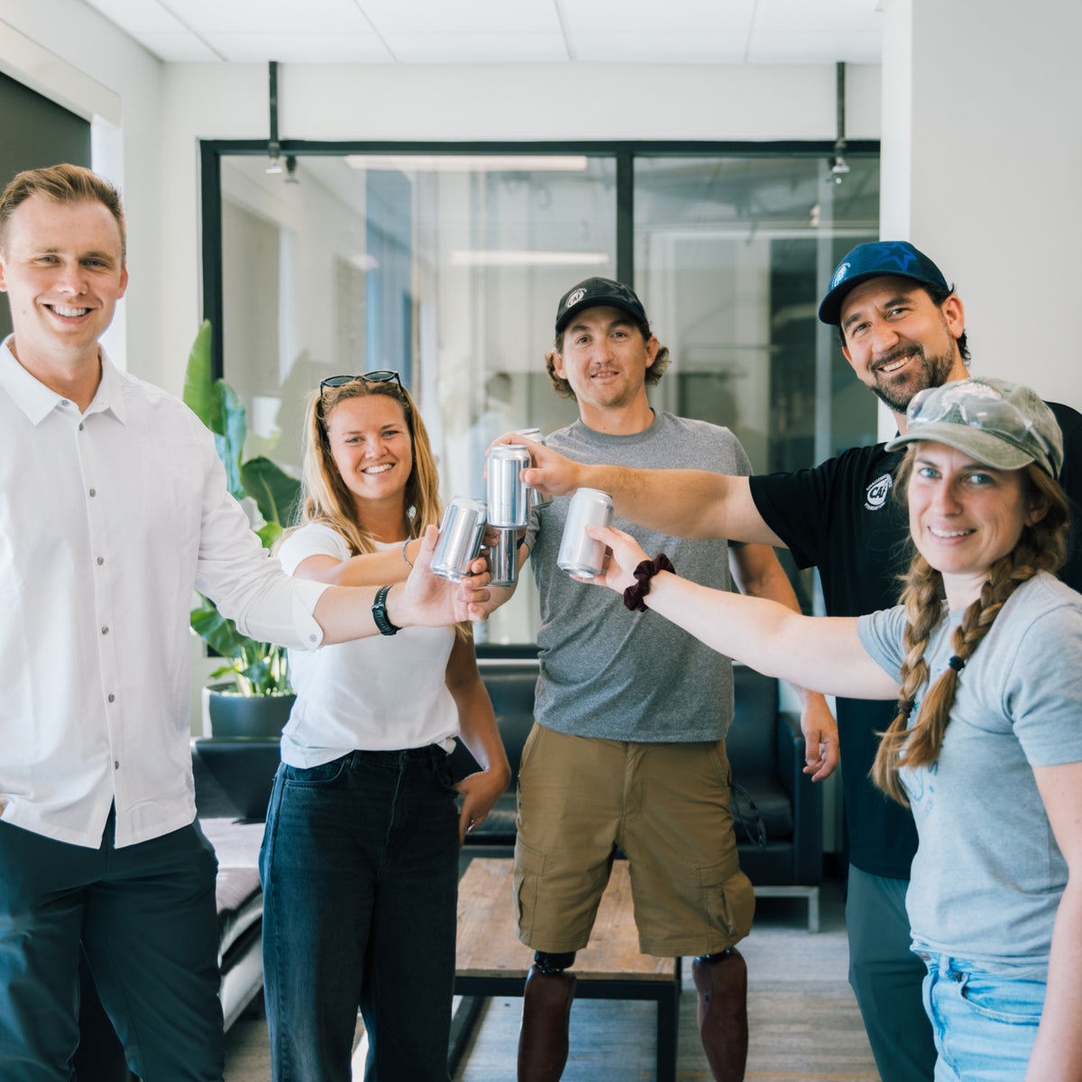 Five people smiling and raising beverage cans together in a bright indoor setting, celebrating community and shared experiences.