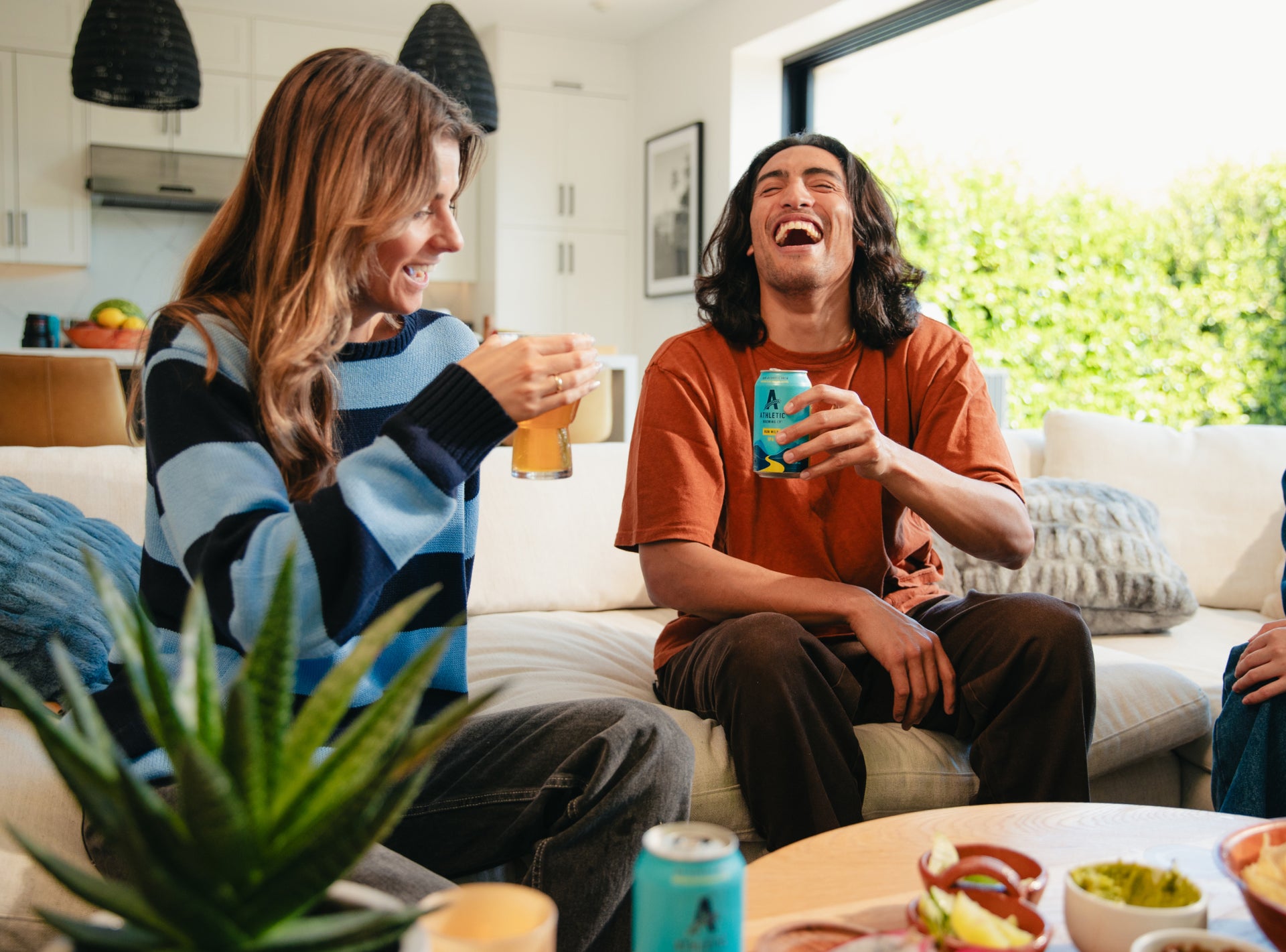 Two people sitting on a couch in a living room, laughing and holding drinks.