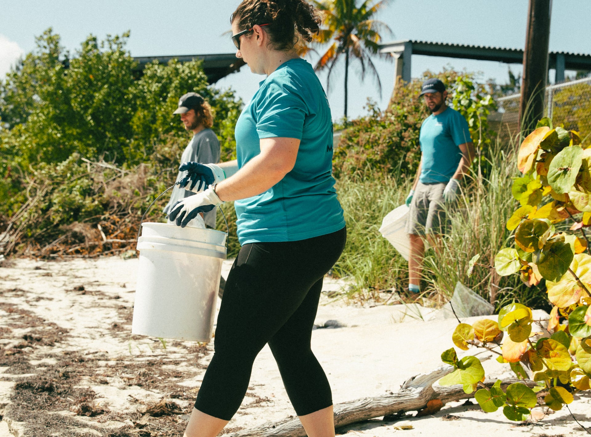 A team of volunteers in teal shirts cleans up a sandy beach. One person in the foreground is carrying a white bucket.